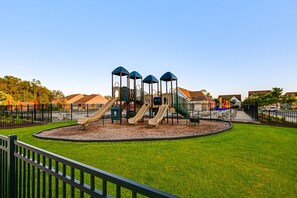 A playground with multiple slides and climbing structures, surrounded by a fenced area and a well-maintained lawn. - Family-Friendly Beach Base with Pool and Shuttle (Ocean View)