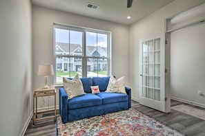 A living room with a blue sofa, patterned rug, and a window with a view of houses. - Family-Friendly Beach Base with Pool and Shuttle (Ocean View)
