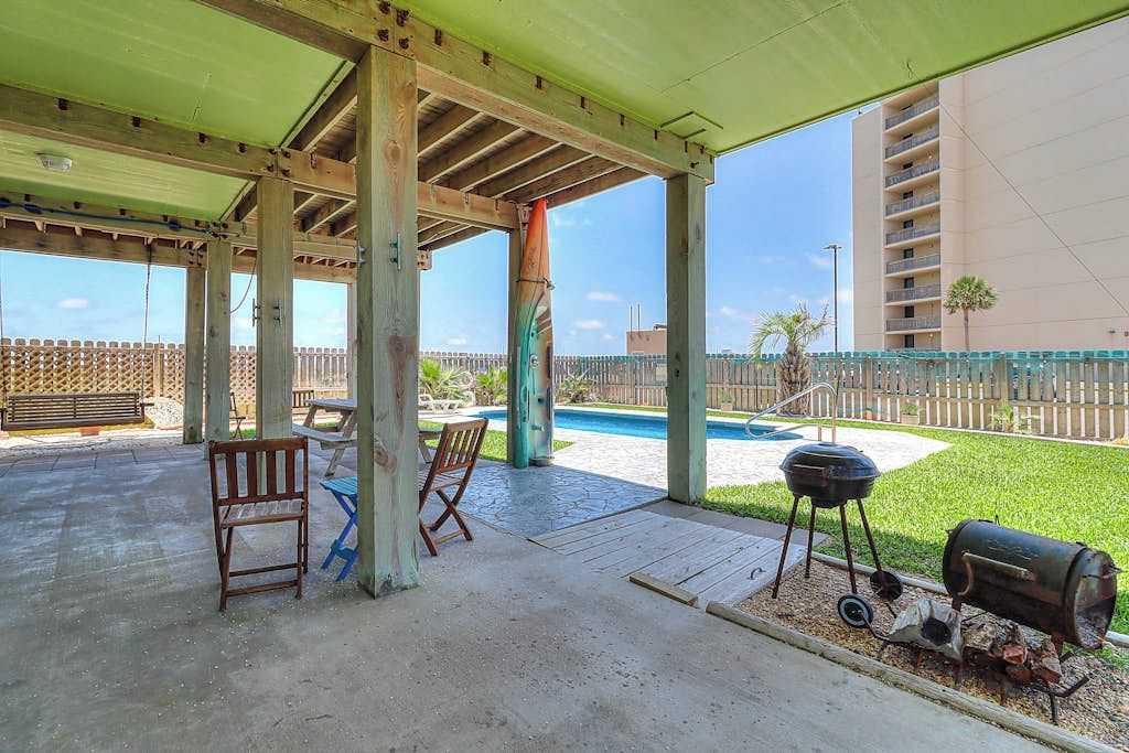 An outdoor patio area with a covered seating area, a barbecue grill, and a pool in the background.