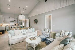 A spacious living area with a white sofa, a round coffee table, and patterned ottomans. A kitchen with modern appliances is visible in the background. - Reserve at St Charles Bay - Cottage By The Sea (Rockport)