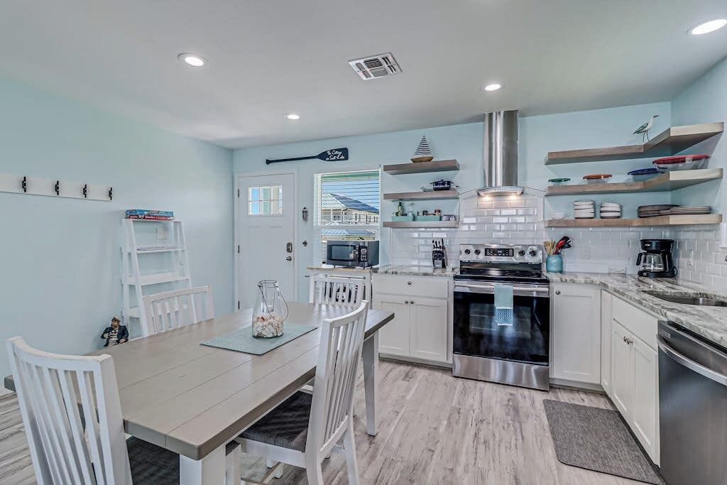 A modern kitchen with white cabinetry, a built-in oven, and a microwave. There is a dining area with a wooden table and white chairs.