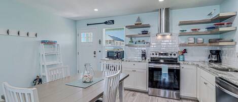 A modern kitchen with white cabinetry, a built-in oven, and a microwave. There is a dining area with a wooden table and white chairs.