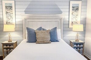 Une chambre à coucher avec un lit blanc, des oreillers bleus à motifs et deux lampes de chevet posées sur des tables de nuit en bois.
