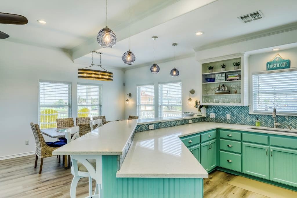A kitchen with a large island, teal cabinetry, pendant lighting, and a view of the outdoors.