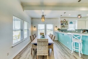 A dining area with a long table and wicker chairs, a kitchen with a teal countertop, and pendant lighting. - Bay View Breeze - Bay Views & Furnished Deck (Rockport)