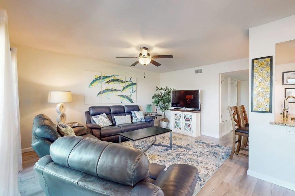 A living room with a sofa, coffee table, and a television. There is a ceiling fan and a plant.