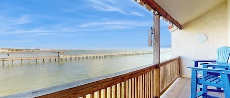 Un balcón con baranda de madera que da a una masa de agua, un muelle que se adentra en el agua y una silla azul.