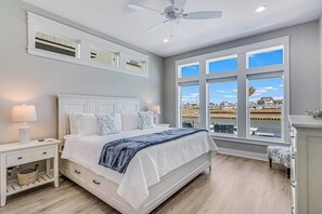 A bedroom with a large bed, a ceiling fan, and a view of a body of water through large windows. - Palm Breeze at the Reserve (Rockport)