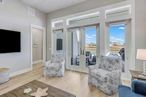 A spacious living room with a large window, two patterned armchairs, a wooden coffee table, and a flat-screen TV mounted on the wall. - Palm Breeze at the Reserve (Rockport)
