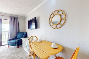 A room with a blue armchair, a wooden table with two chairs, a television mounted on the wall, and a decorative mirror. - Casa Del Mar - The Seaside (Galveston)