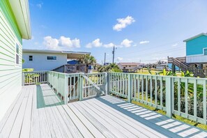 A wooden deck with a railing, leading to a building with a blue exterior. - The Treehouse - Steps to the Beach & Sunny Deck (Galveston)