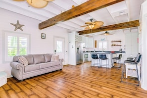 A spacious living area with a wooden ceiling, a gray sofa, and a kitchen in the background. - The Treehouse - Steps to the Beach & Sunny Deck (Galveston)