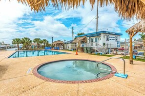 A swimming pool area with a hot tub, a building with a blue roof, and palm trees. - Pink Lady by the Sea (Galveston)
