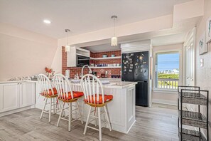 A kitchen with white cabinets, a brick backsplash, a black refrigerator, and a dining area with white chairs and orange cushions. - Pink Lady by the Sea (Galveston)