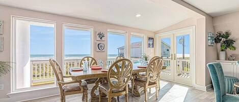 A dining area with a large table and chairs, large windows with a view of the ocean, and a balcony with a railing.