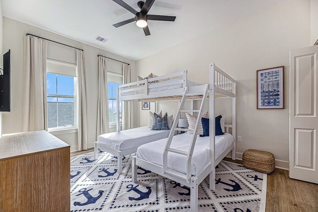 A room with bunk beds, a ceiling fan, a window with curtains, and a framed picture on the wall.