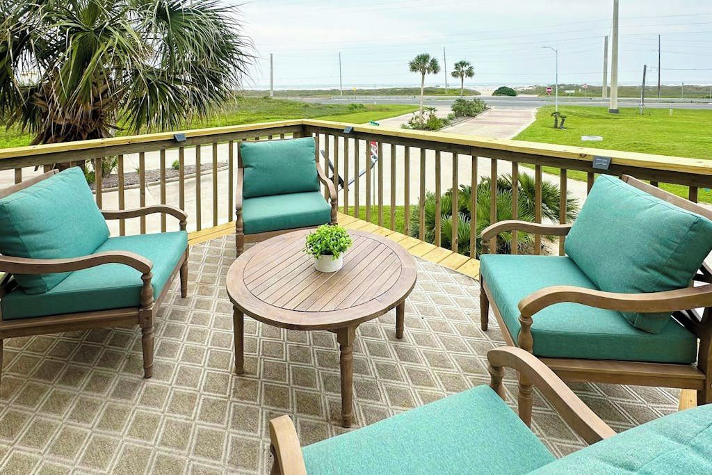 A covered outdoor area with a wooden deck, two green cushioned chairs, a round wooden table with a plant, and a view of a road and greenery.