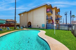 A swimming pool with lounge chairs, a building with balconies, and a fenced area. - Marlin Arms Condominiums #9 (South Padre Island)