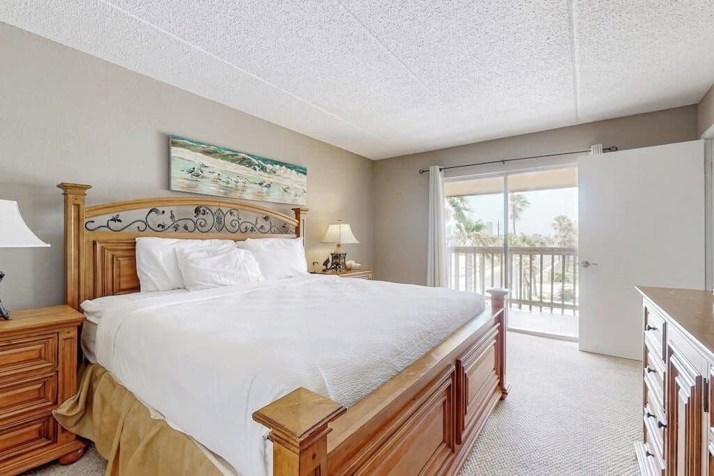 A bedroom with a large bed, wooden headboard, a nightstand with a lamp, a dresser, and a view of the outdoors through a sliding glass door.