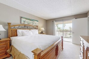 Quarto com cama grande, cabeceira de madeira, mesa de cabeceira com abajur, cômoda e vista para o exterior por uma porta de vidro deslizante.