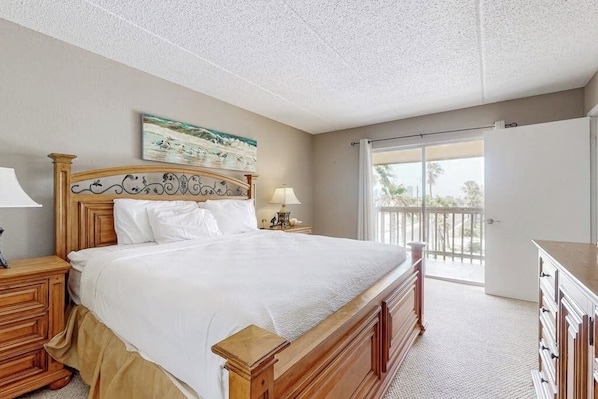 A bedroom with a large bed, wooden headboard, a nightstand with a lamp, a dresser, and a view of the outdoors through a sliding glass door. - Marlin Arms Condominiums #9 (South Padre Island)