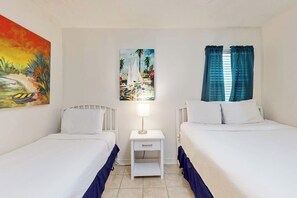 Une chambre d’hôtel avec deux lits, une table de chevet, un tableau représentant un voilier et une fenêtre avec des rideaux.