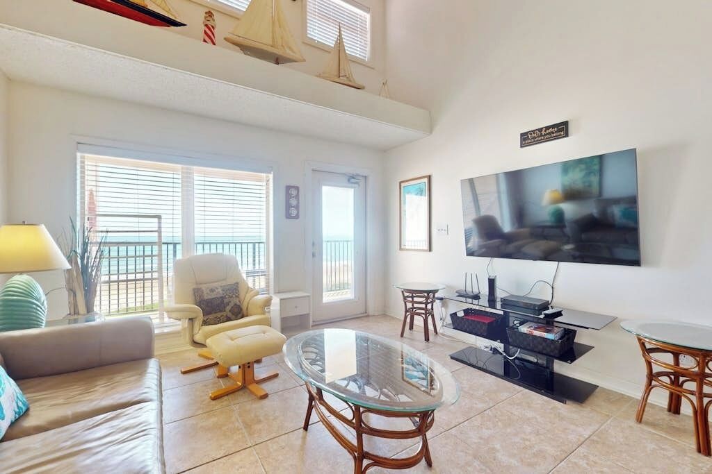 A living room with a sofa, armchair, glass coffee table, TV, and a small table with electronic devices.