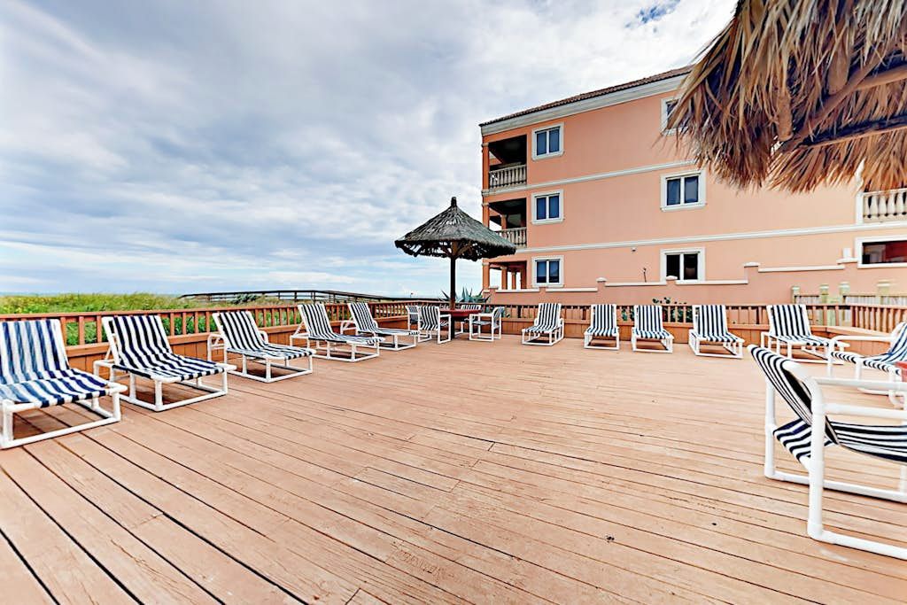 A deck with lounge chairs and a thatched umbrella, overlooking a building with balconies.