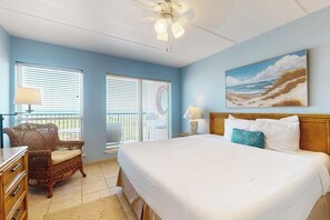A bedroom with a bed, two windows, a ceiling fan, a painting of a beach, and a wicker chair. - Suntide II Condominiums #207 (South Padre Island)