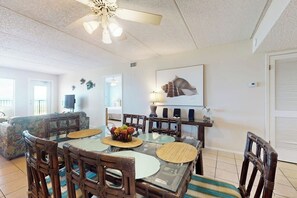 A dining area with a glass-top table, wooden chairs, a ceiling fan, and a wall-mounted TV. - Suntide II Condominiums #207 (South Padre Island)