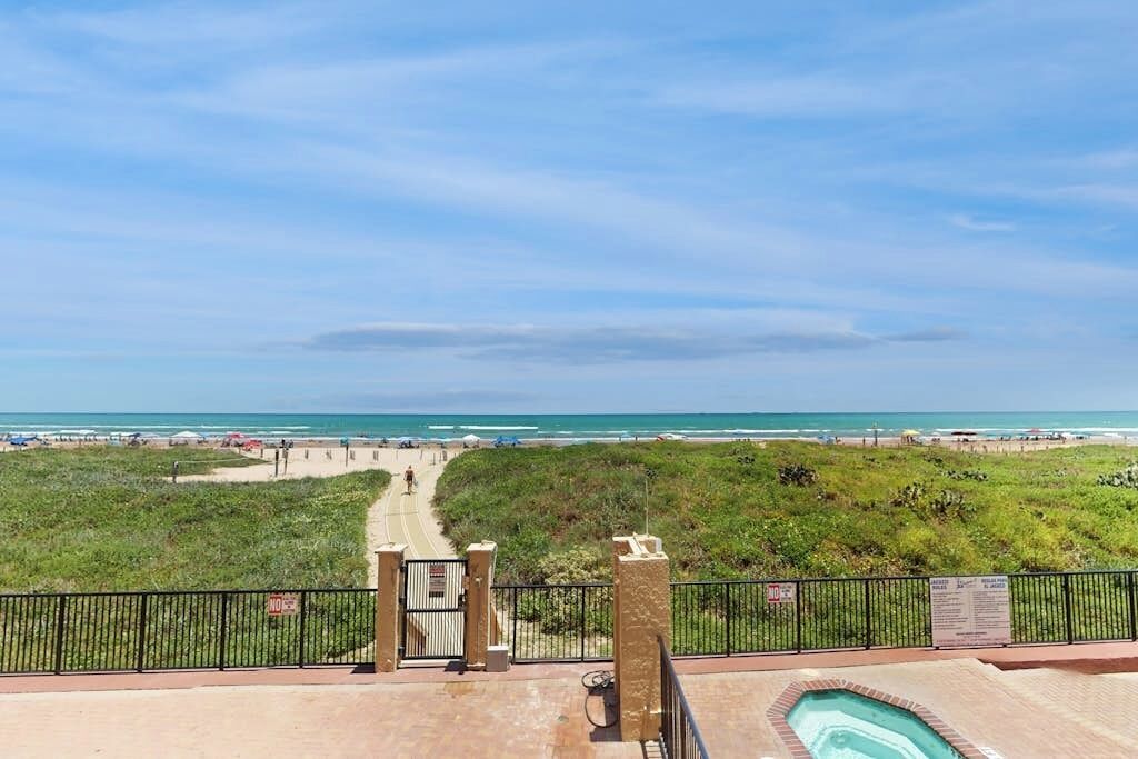 A view of a beach with a pathway leading to the water, a pool area with a fence, and a clear sky.