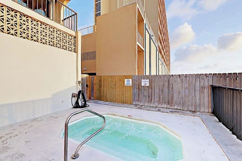A swimming pool with a wooden fence, a metal pool ladder, and a building with balconies in the background.