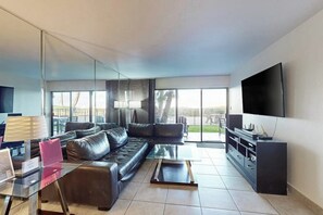 A modern living room with a sectional sofa, a glass coffee table, a flat-screen TV, and a large window offering a view of the outdoors. - Ocean Vista Condominiums #102 (South Padre Island)