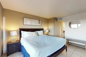 Une chambre d’hôtel avec un grand lit, des tables de chevet avec des lampes, un banc, un miroir fixé au mur et une porte.