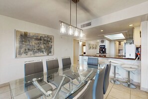A modern dining area with a glass table and chairs, a hanging light fixture, and a large abstract painting on the wall. - Ocean Vista Condominiums #102 (South Padre Island)