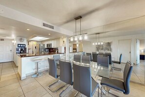 A modern kitchen with a central island, bar stools, and a dining area with a glass table and chairs. - Ocean Vista Condominiums #102 (South Padre Island)