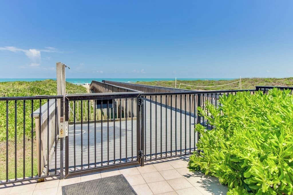 A gated entrance to a beach area with a clear view of the ocean and a pathway leading to a building.