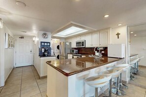 A modern kitchen with a central island, bar stools, and a cross on the wall. - Ocean Vista Condominiums #102 (South Padre Island)
