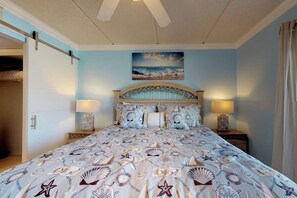 A bedroom with a bed, two bedside tables, a headboard, a wall-mounted painting, and a ceiling fan. - Beachview Condominiums #204 (South Padre Island)