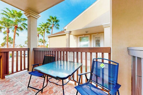An outdoor patio with a table and chairs, palm trees, and a building in the background. - Escape Condominiums #7 (South Padre Island)