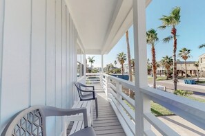 A balcony with white railings, a row of chairs, and a view of palm trees and buildings. - Lazy Seagull Beach House (South Padre Island)
