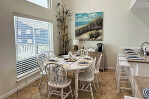 A dining area with a round table, white chairs, a painting of a beach scene, and a window with blinds. - Seabreeze Serenity - Beachfront & Dog-Friendly (Galveston)