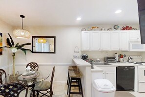 A compact kitchen with white cabinets, a small dining area with wicker chairs, and a white trash bin. - La Internacional Condominiums #210 (South Padre Island)