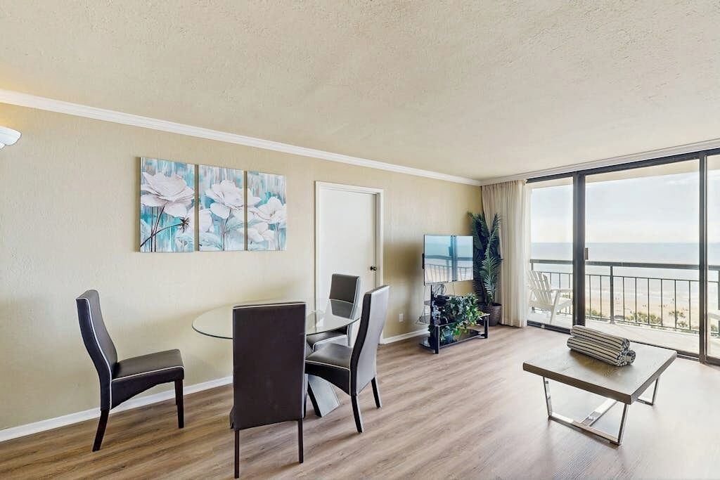 Een kamer met een eettafel, stoelen, kunst aan de muur, een deur en een groot raam met uitzicht.