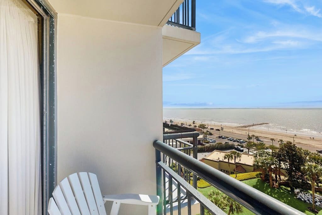 A balcony with a white chair, a view of the beach, and a clear sky.
