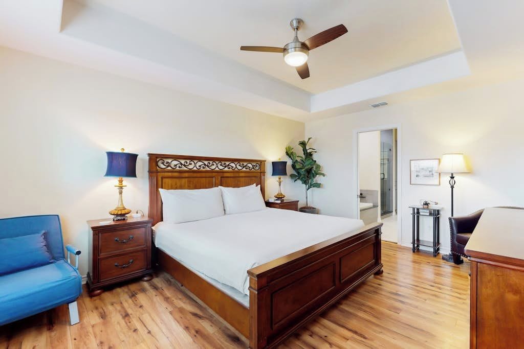 A bedroom with a wooden bed, a ceiling fan, a nightstand with a lamp, a blue chair, and a small table with a lamp.