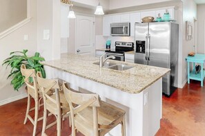 A kitchen with white cabinets, stainless steel appliances, a granite countertop, and a bar stool area. - Nemo Cay - Too The Beach (Corpus Christi)