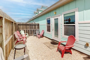A patio area with Adirondack chairs and a small table, a wooden fence, and a building with a light blue exterior. - Nemo Cay - Too The Beach (Corpus Christi)