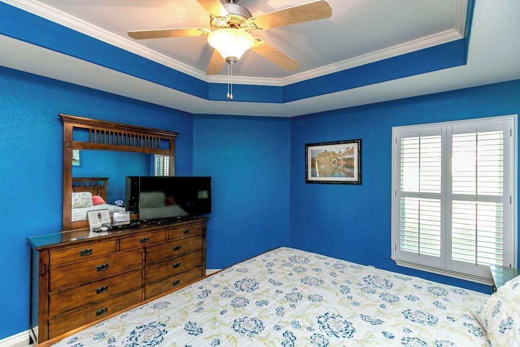 Een slaapkamer met een bed, een dressoir, een televisie, een plafondventilator, een raam met jaloezieën en een lijst met foto aan de muur.