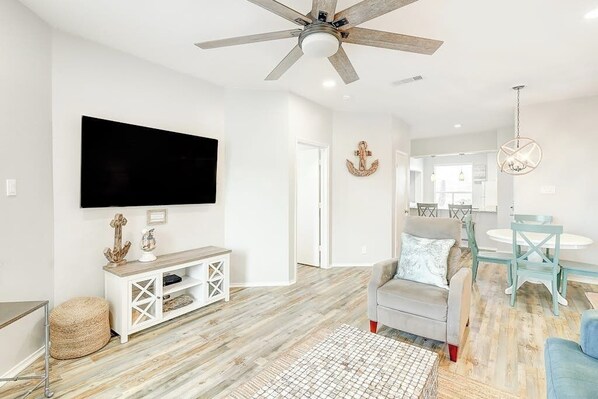 En moderne stue med loftventilator, et fladskærms-tv, en lysegrå sofa, et rundt sofabord og et spiseområde med et hvidt bord og blå stole.
